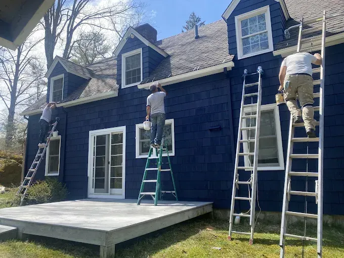 Three people paint the exterior of a blue house with white trim; ladders are in use.