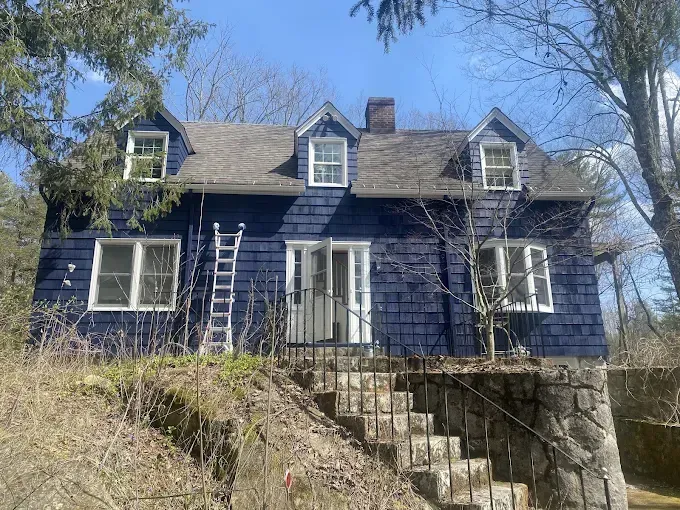Blue shingled house with white trim and three dormers, built on a stone foundation with outdoor stairs.
