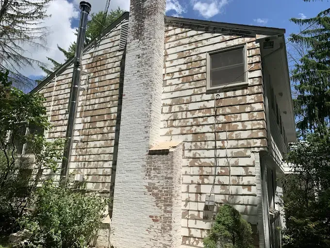 Side of a house with weathered, peeling white shingles and a brick chimney.