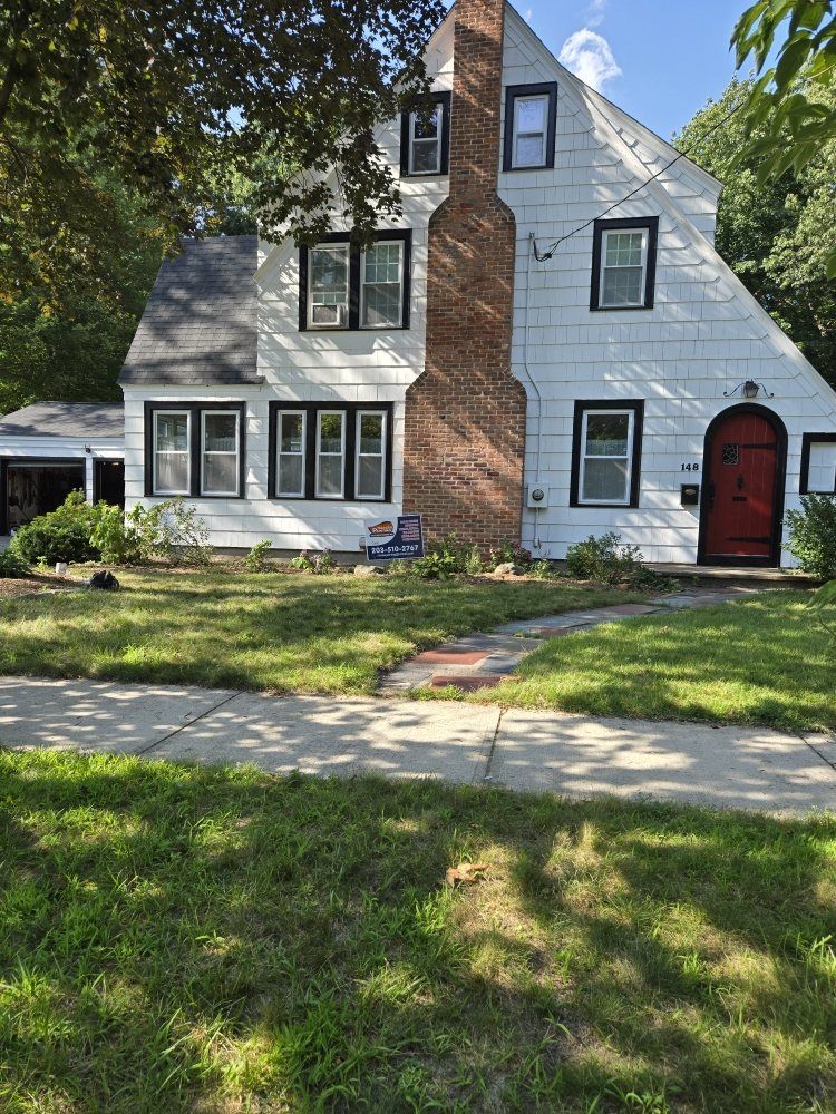 White house with black-trimmed windows, a red door, and a brick chimney, set on a green lawn.