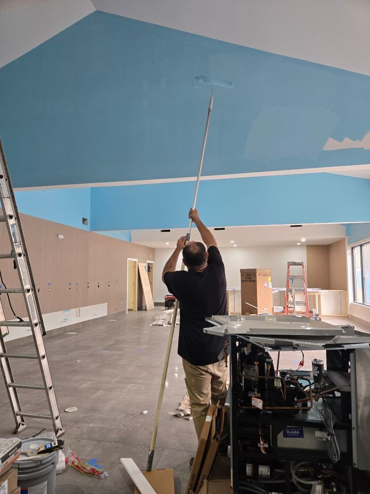 Person paints a light blue ceiling with a roller on a long handle in an unfinished room.