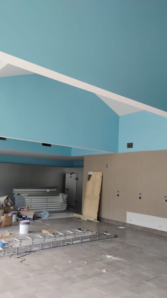 Construction site interior, light blue ceiling, beige wall, ladder, and supplies scattered on a concrete floor.
