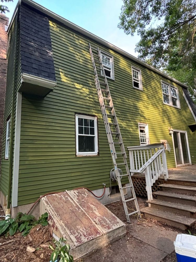 Green house exterior with a ladder against the side, a brick chimney, and a wooden deck.