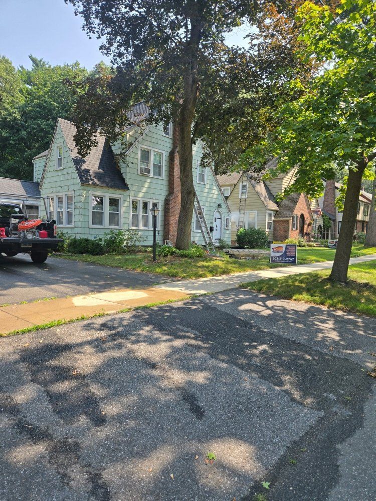 Light green house with black roof, large tree, and parked truck on a sunny day.