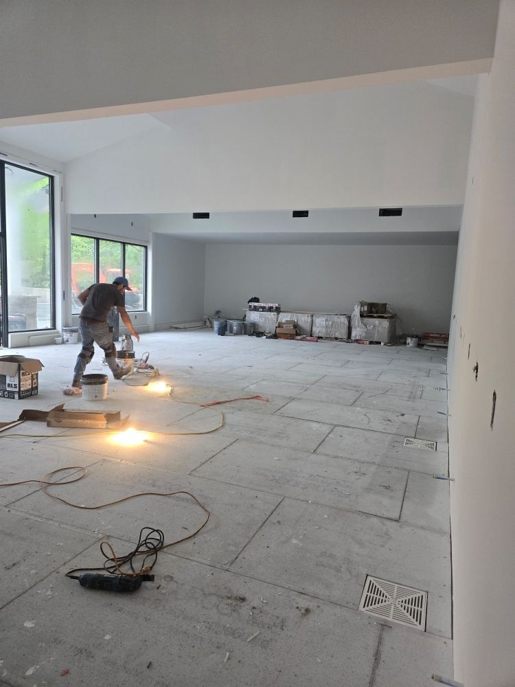 Man using a floor grinder in a large room, tiles on floor, windows and white walls.