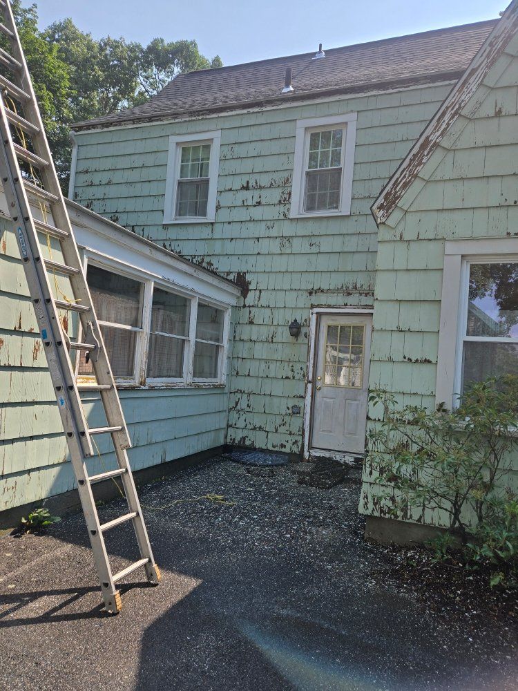 Back of a two-story teal house with peeling paint, a ladder, and a gravel patio on a sunny day.