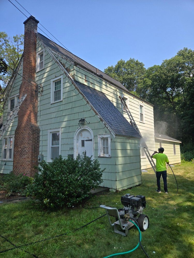Person power washing a two-story green house with a brick chimney. Pressure washer and ladder visible. Sunny day.