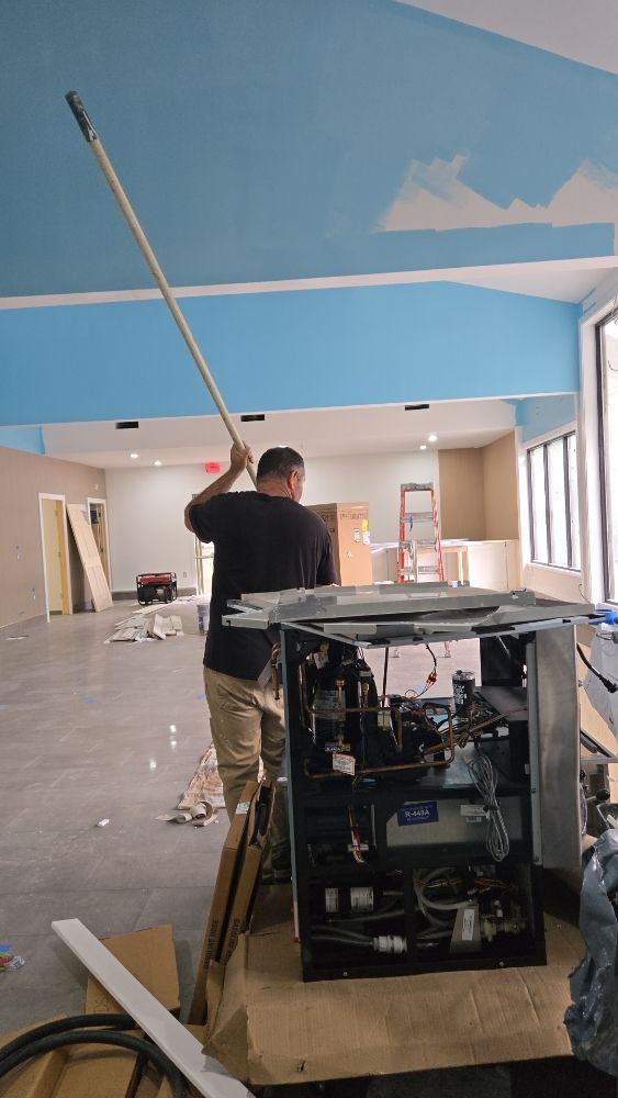 Man uses a pole to work on a blue ceiling in a room under construction. An open machine sits nearby.
