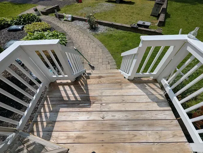 Wooden stairs with white railings lead down to a paved pathway in a yard with grass and landscaping.