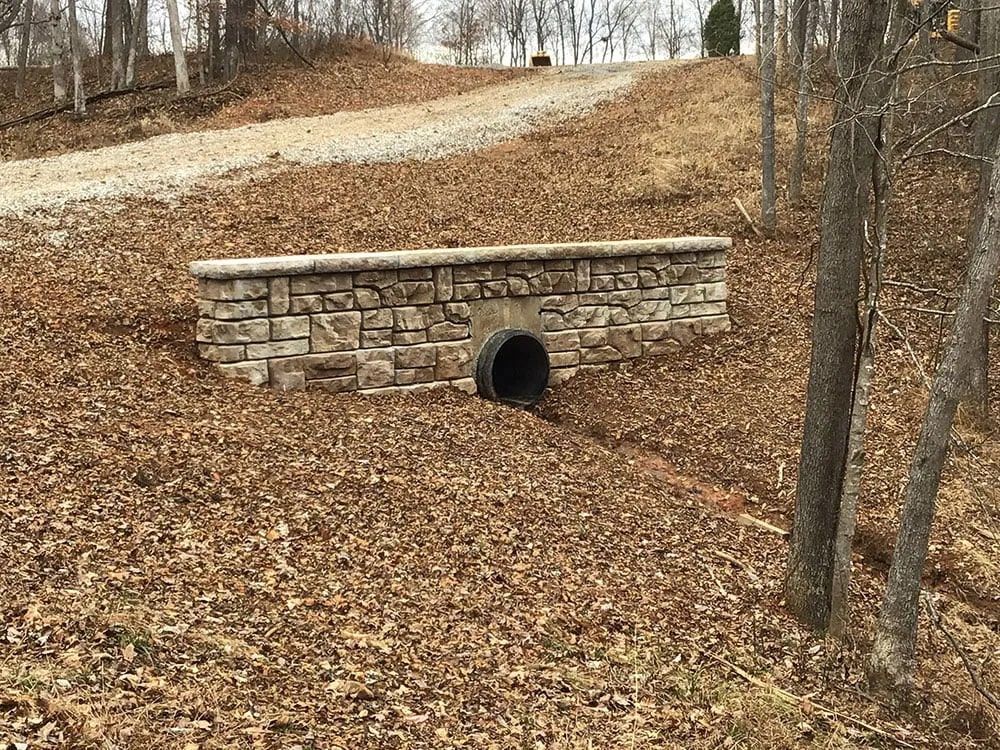 A stone wall with a pipe going through it in the middle of a forest.