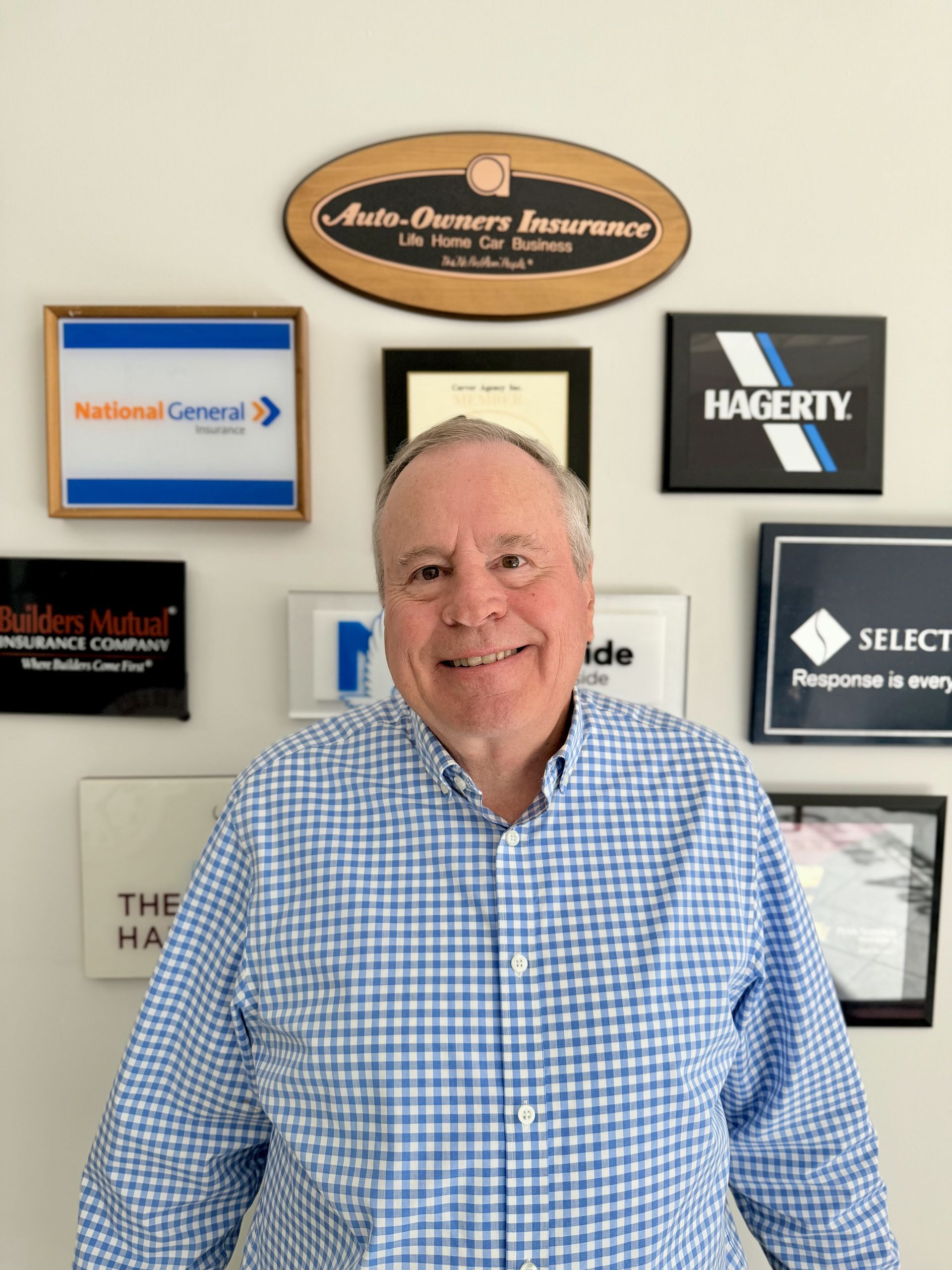 A smiling person in a blue and white checkered shirt standing in front of a wall decorated with insurance company logos.