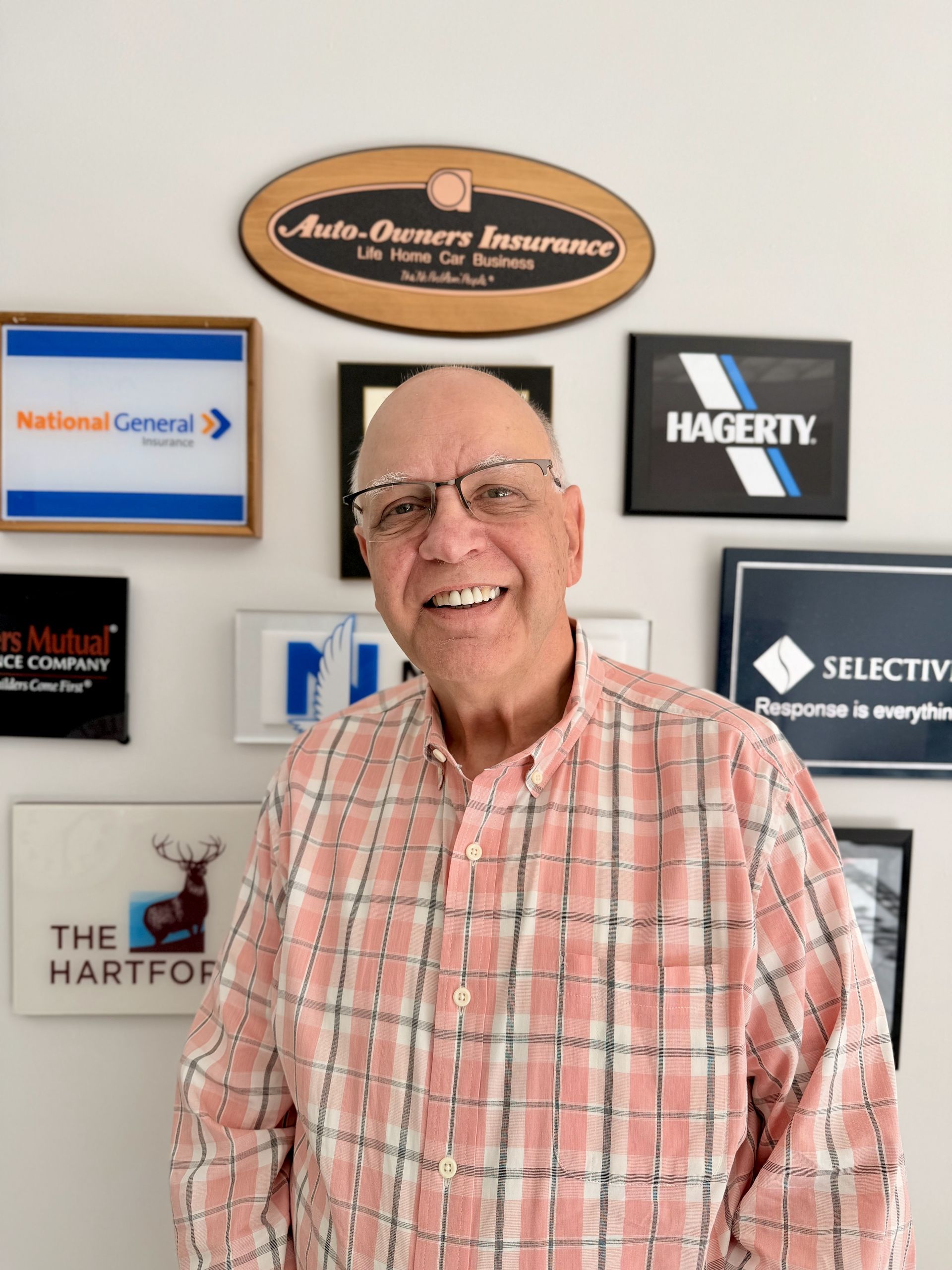 A smiling individual in a pink plaid shirt stands in front of a wall featuring various insurance company logos.