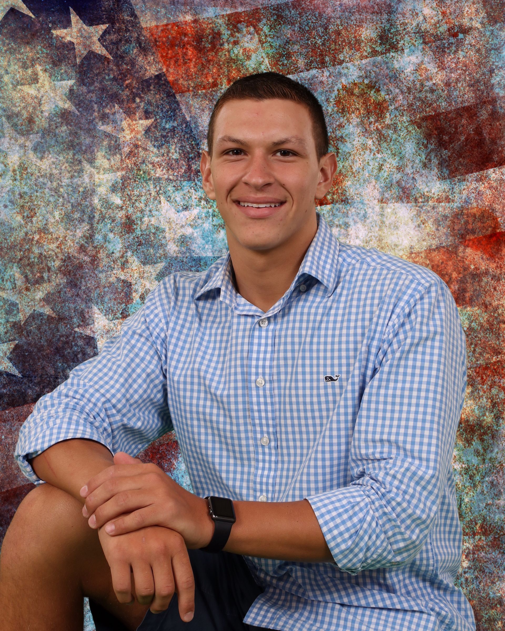 A man in a blue plaid shirt is sitting in front of an american flag.