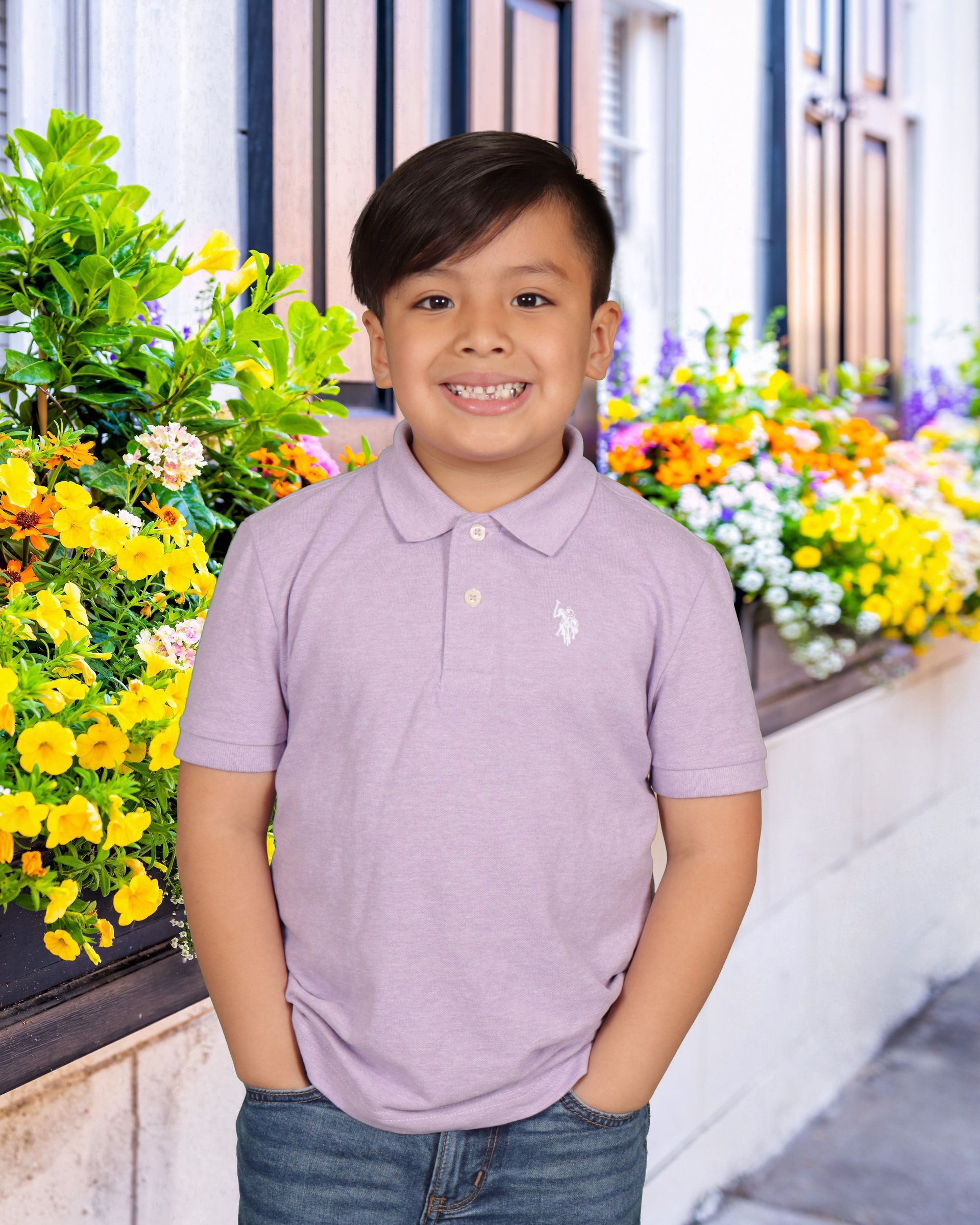 A young boy wearing a purple polo shirt is standing in front of flowers.