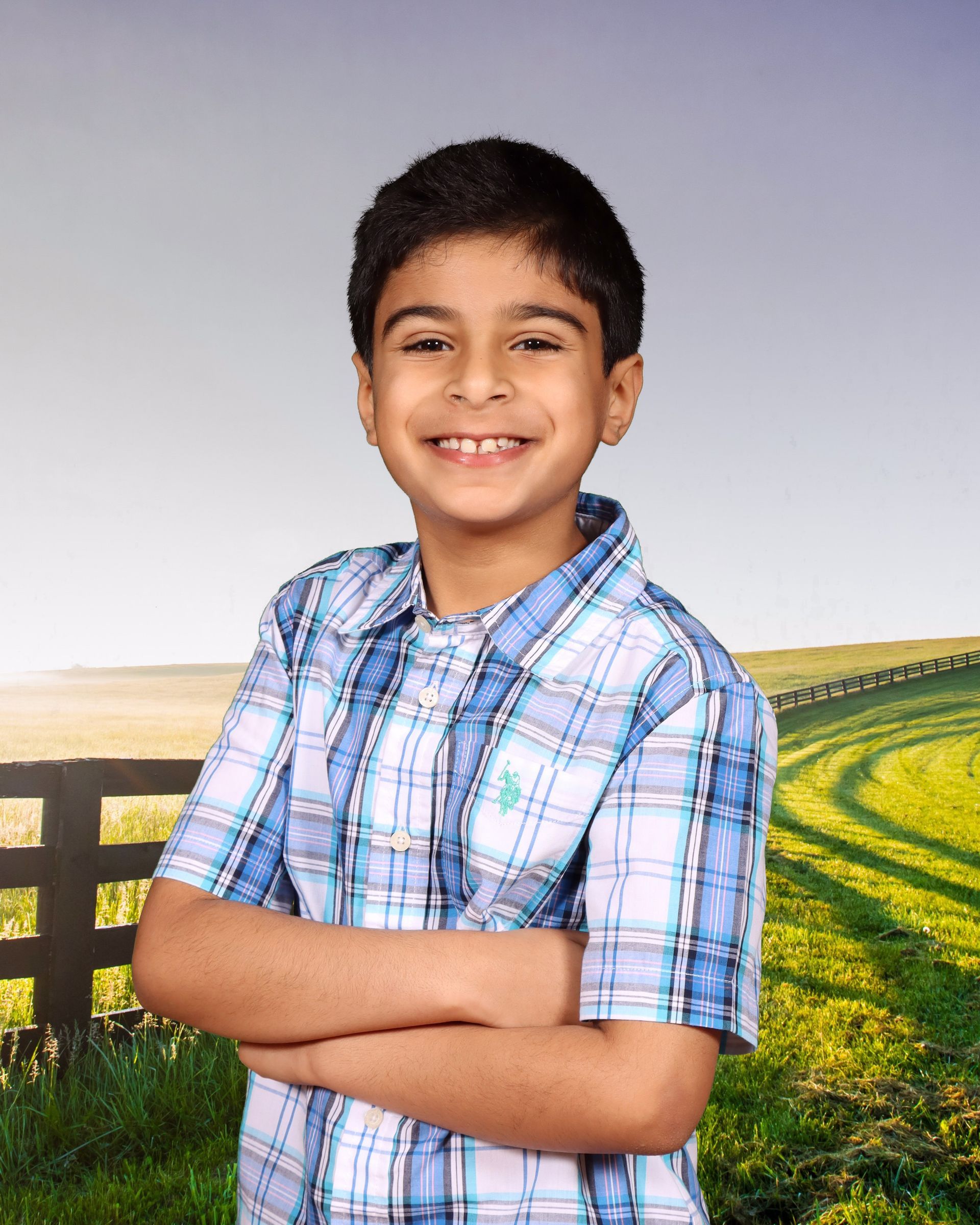 A young boy in a plaid shirt stands with his arms crossed in front of a field