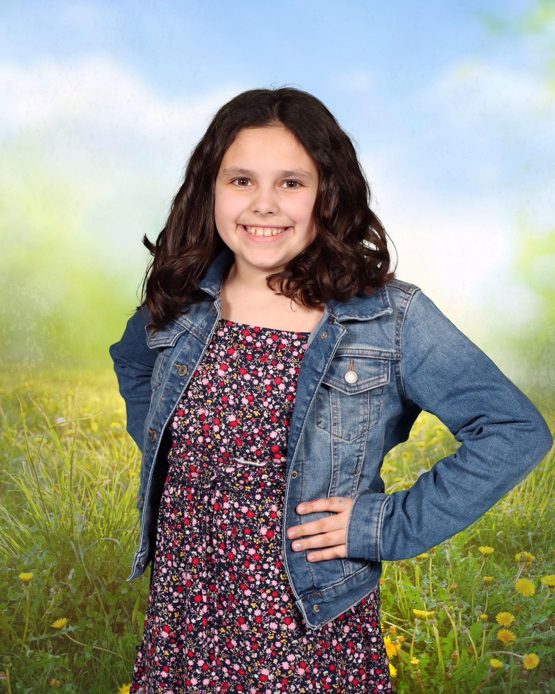 A young girl wearing a denim jacket and a dress is standing in a field.