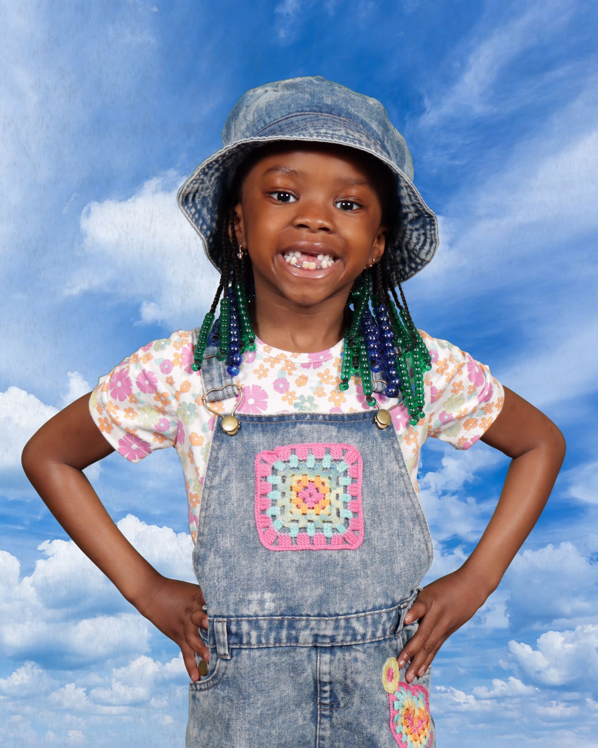 A little girl wearing overalls and a hat is standing with her hands on her hips