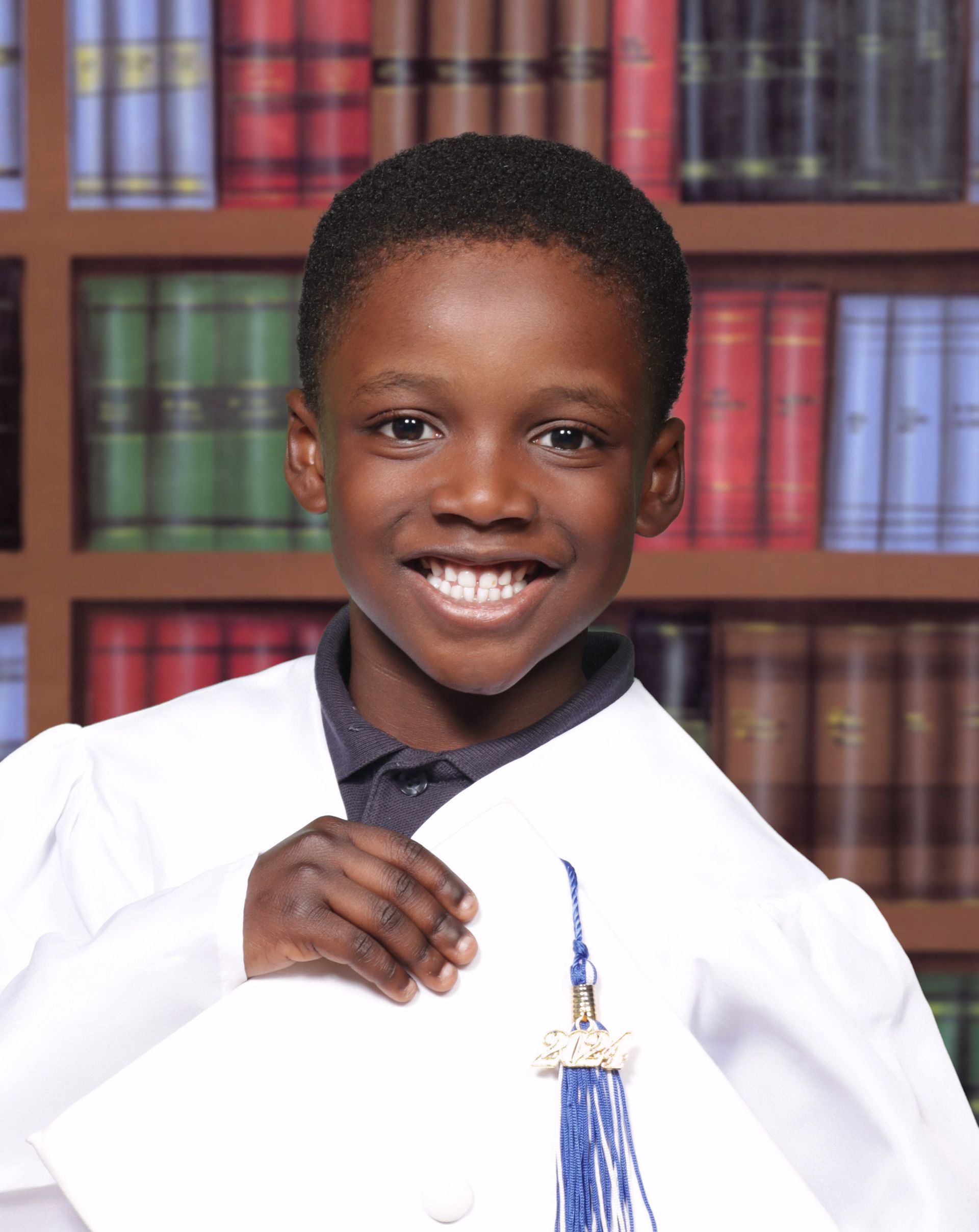 A young boy in a graduation cap and gown holds a diploma in front of a bookshelf.