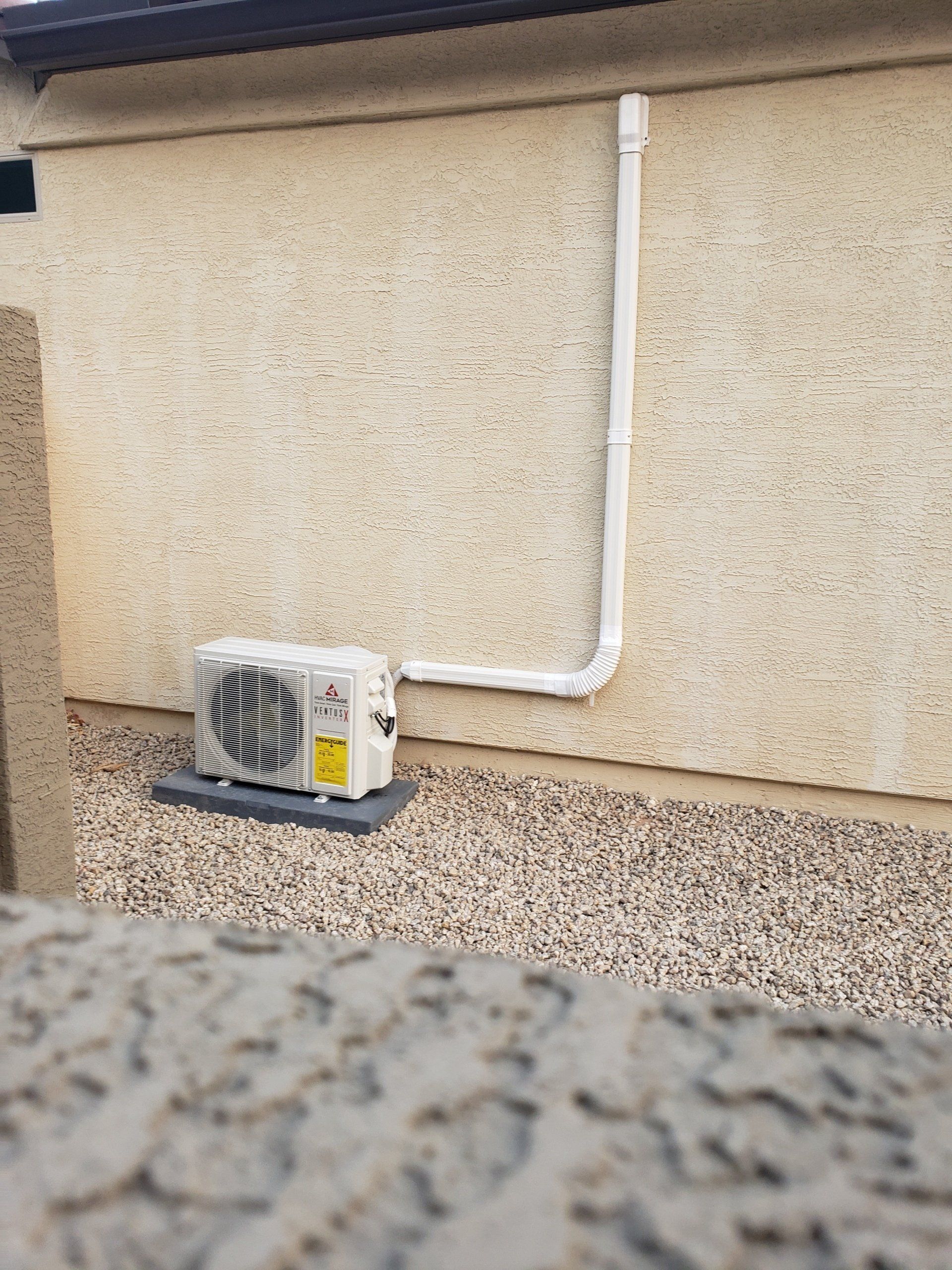 An outdoor air conditioning unit next to a stucco wall, with piping. Gravel ground.