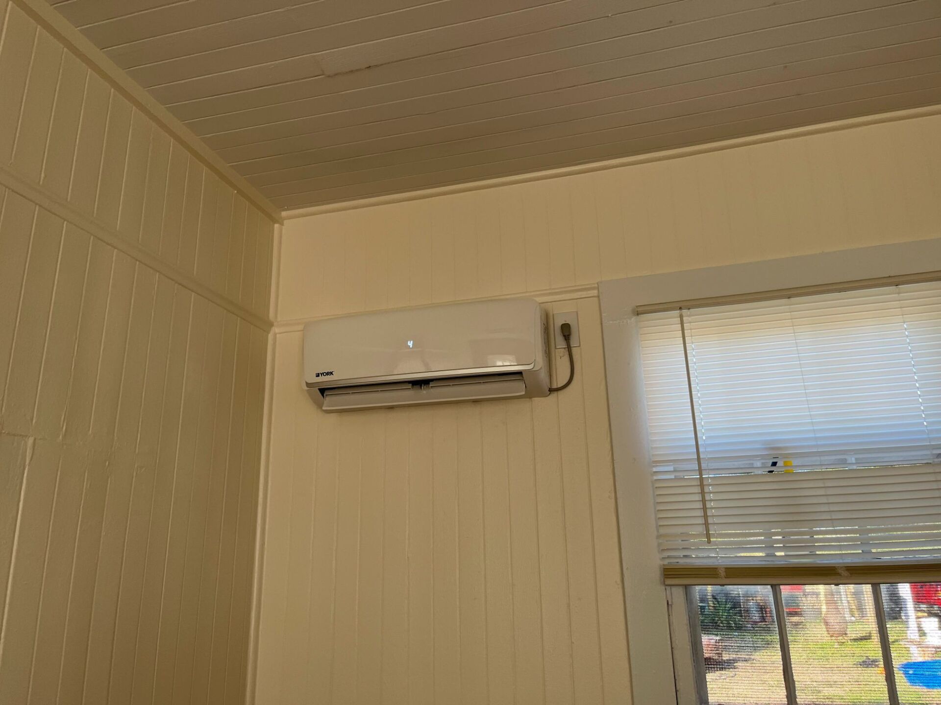 An air conditioning unit mounted on a beige wall inside a room with a window and a ceiling.
