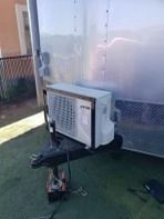 An air conditioner mounted on a trailer hitch. Trailer is silver, on green grass.