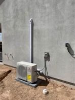 Air conditioning unit mounted on a concrete slab against a light gray stucco wall; tubing ascends.