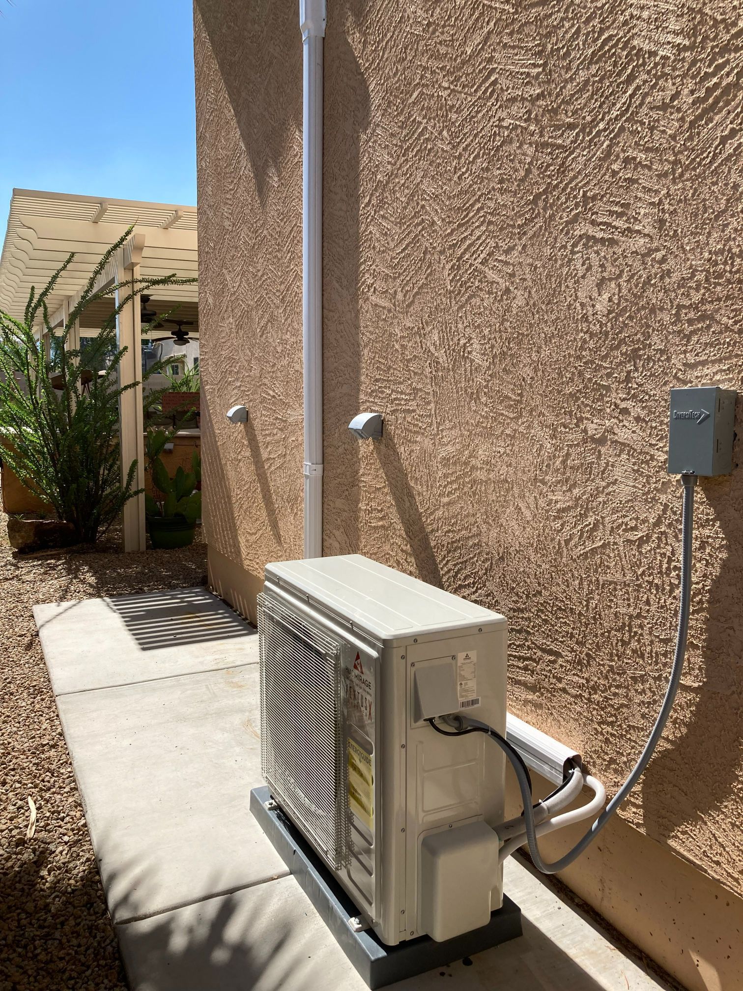 Air conditioning unit mounted on a concrete slab next to a stucco wall, connected to electrical box by a metal conduit.