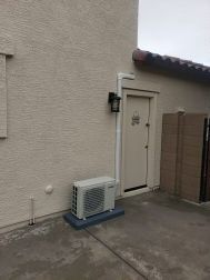 Exterior view: Air conditioning unit on a concrete pad next to a door and light fixture on a stucco wall.