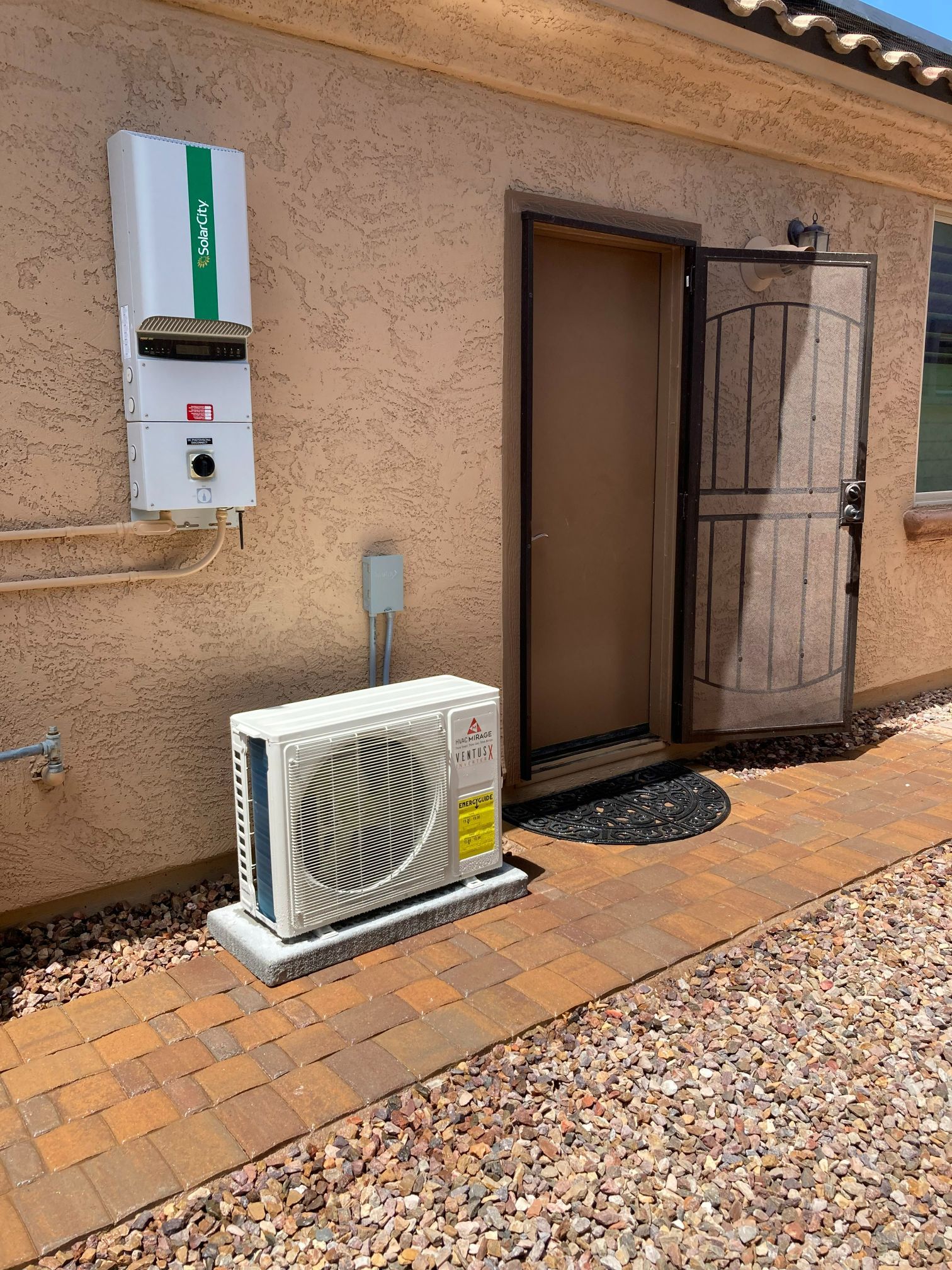 Outdoor electrical equipment next to a door. A white solar inverter and air conditioning unit sit on brick and concrete.