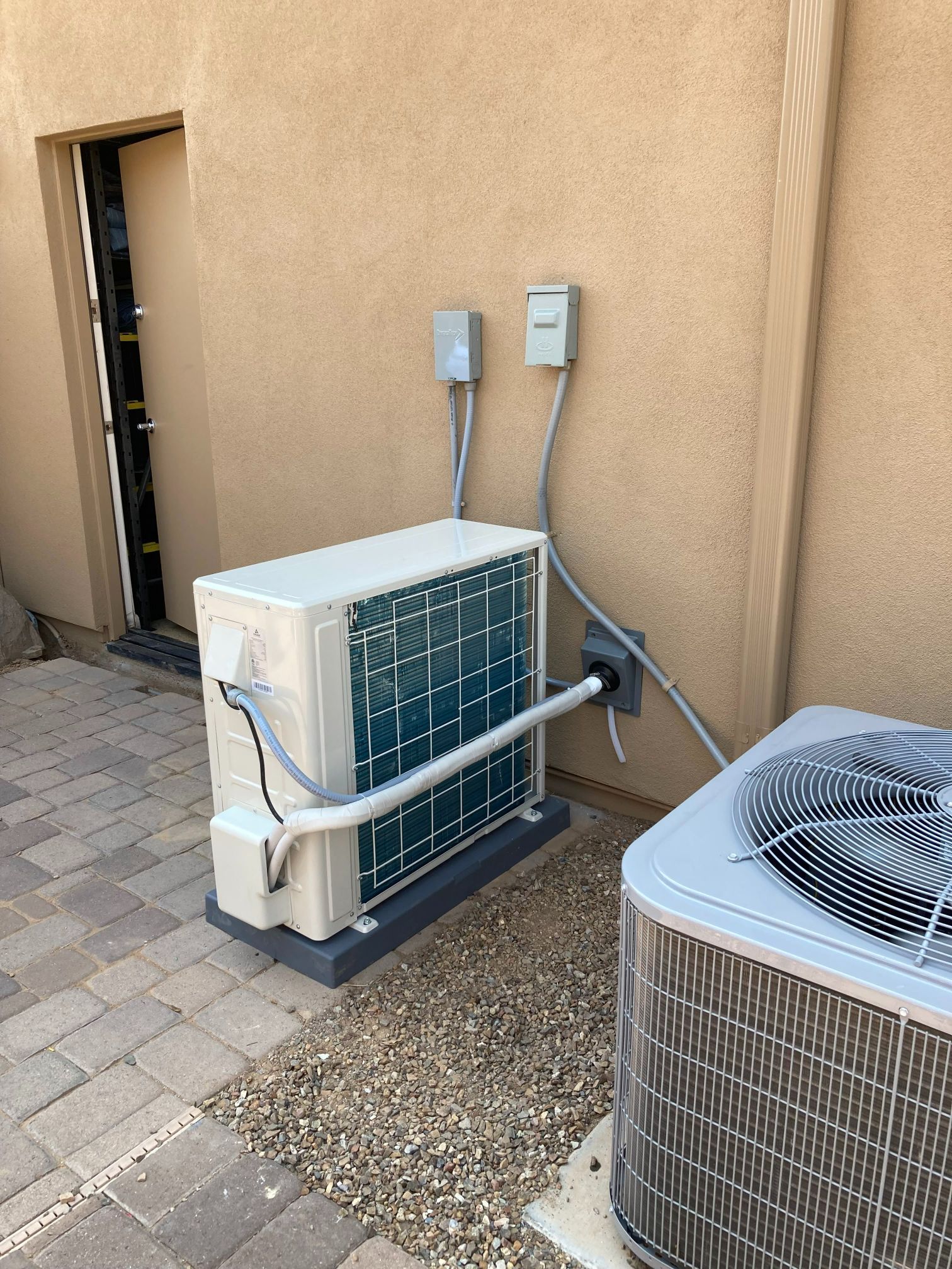 HVAC units mounted on concrete pads beside an exterior wall with electrical boxes and pipes.