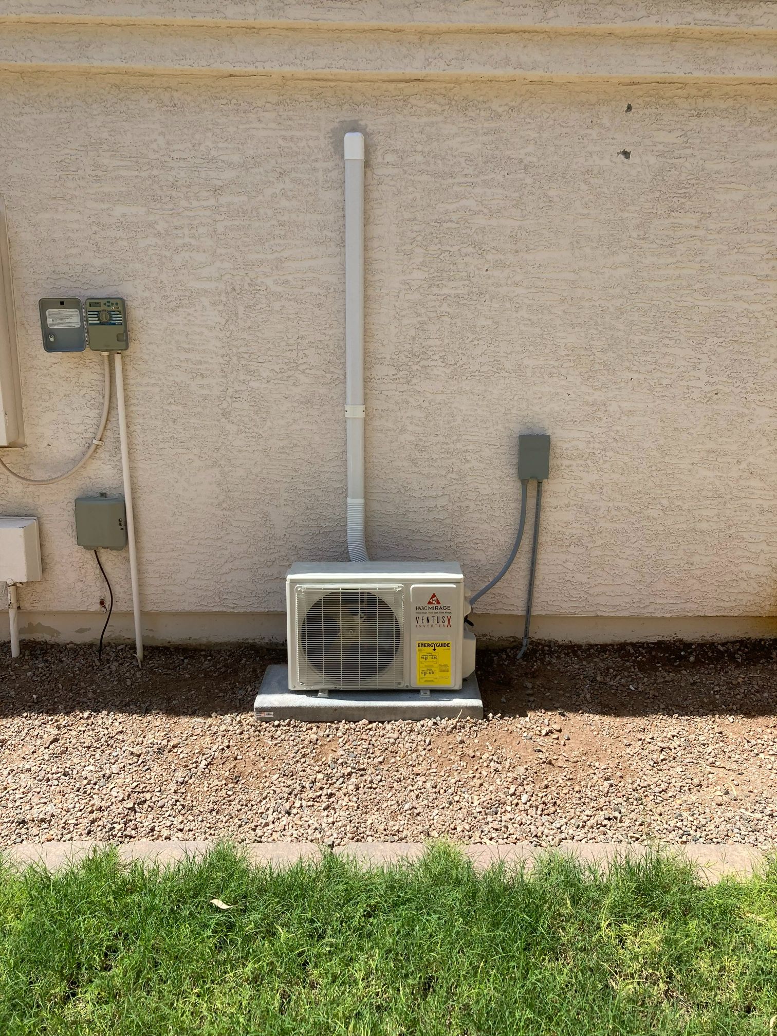 A small metal HVAC unit on a concrete pad near a beige stucco wall with pipes and electrical boxes.