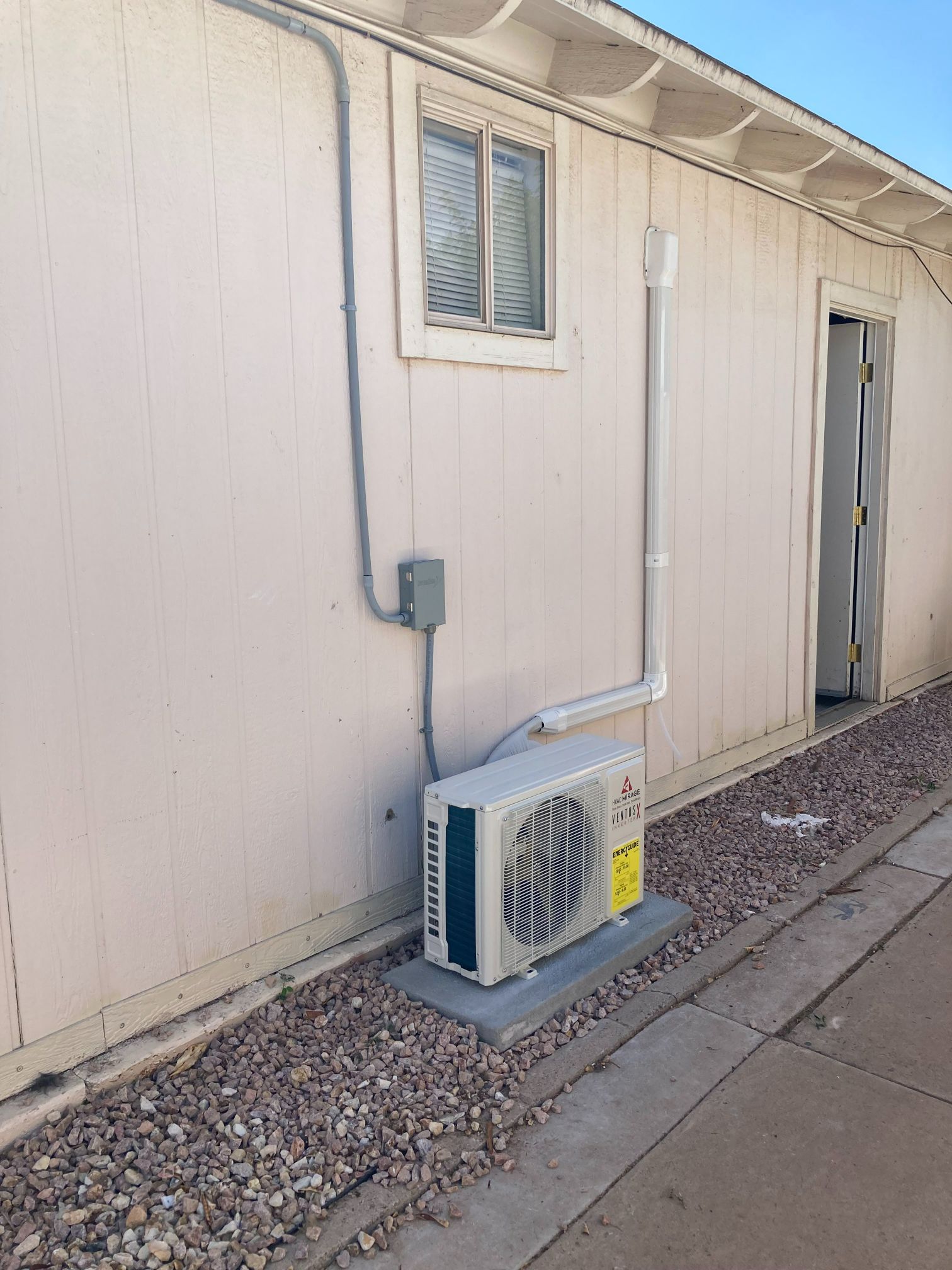 Mini-split AC unit mounted on a concrete pad outside a building with electrical conduit and a window.