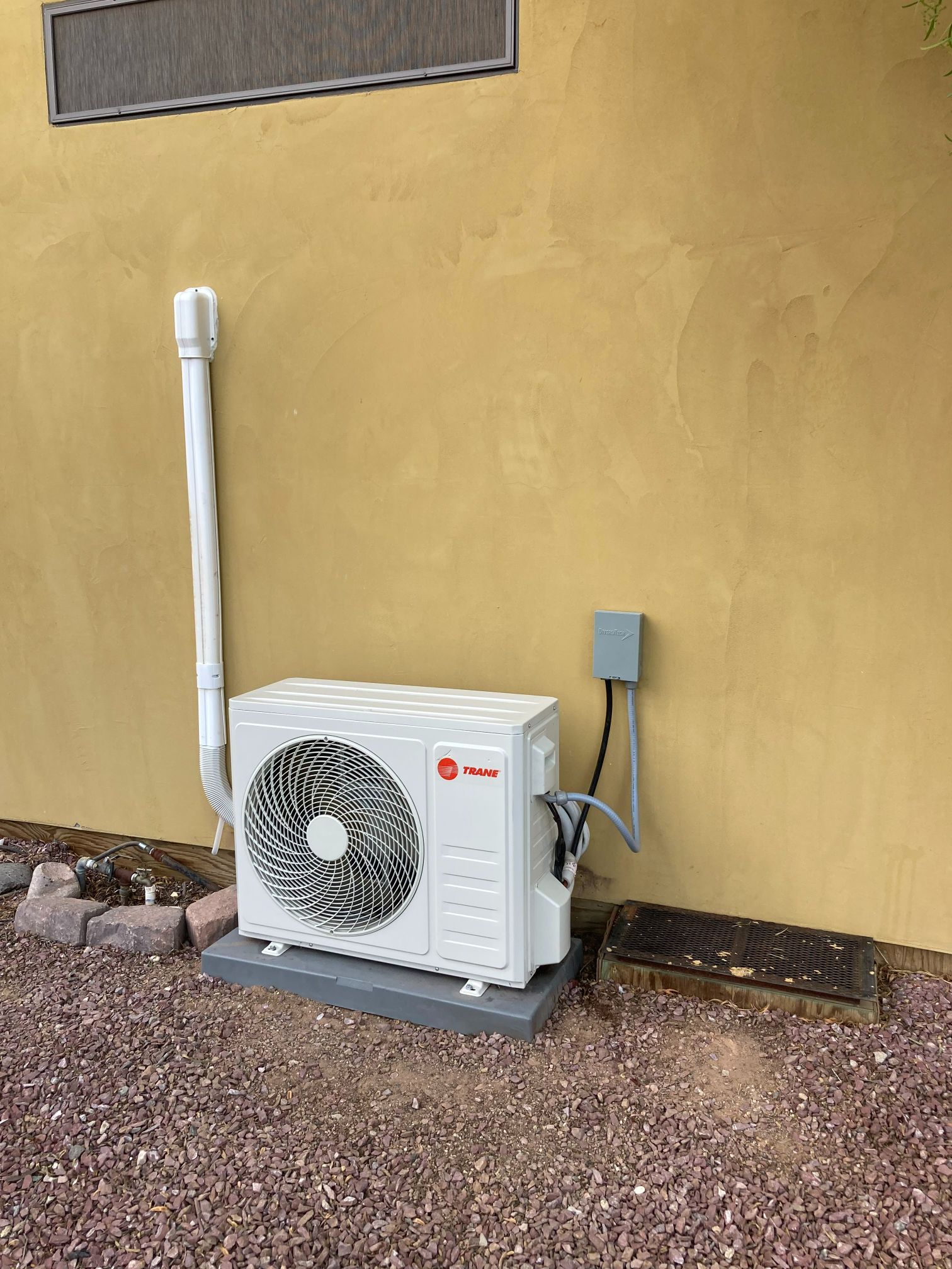 Exterior view of a white air conditioning unit against a yellow stucco wall, with a pipe and electrical box.