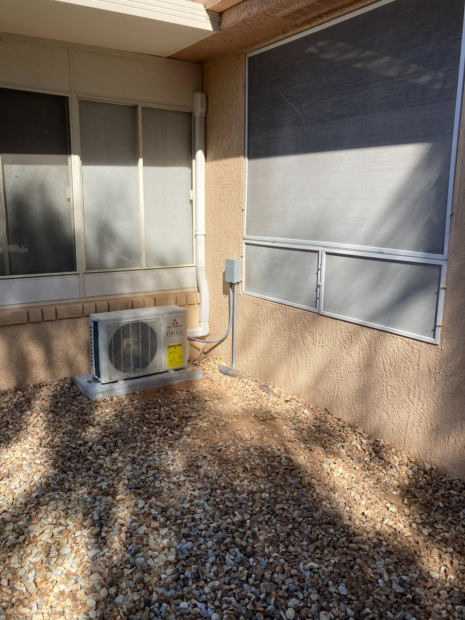 Exterior air conditioning unit next to a wall with windows, on a gravel surface.