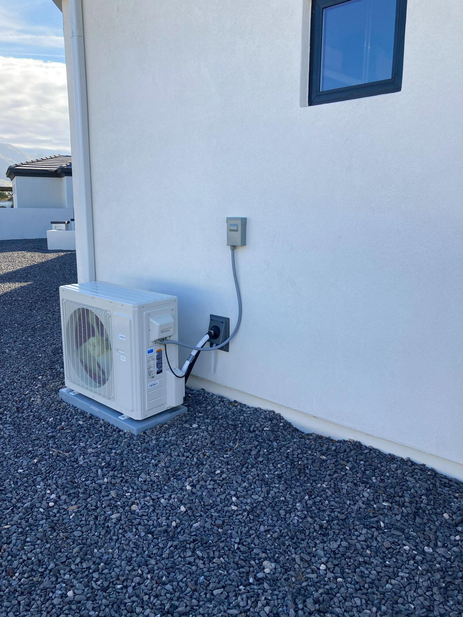 White air conditioning unit mounted on gravel outside a white building with a gray electrical outlet.