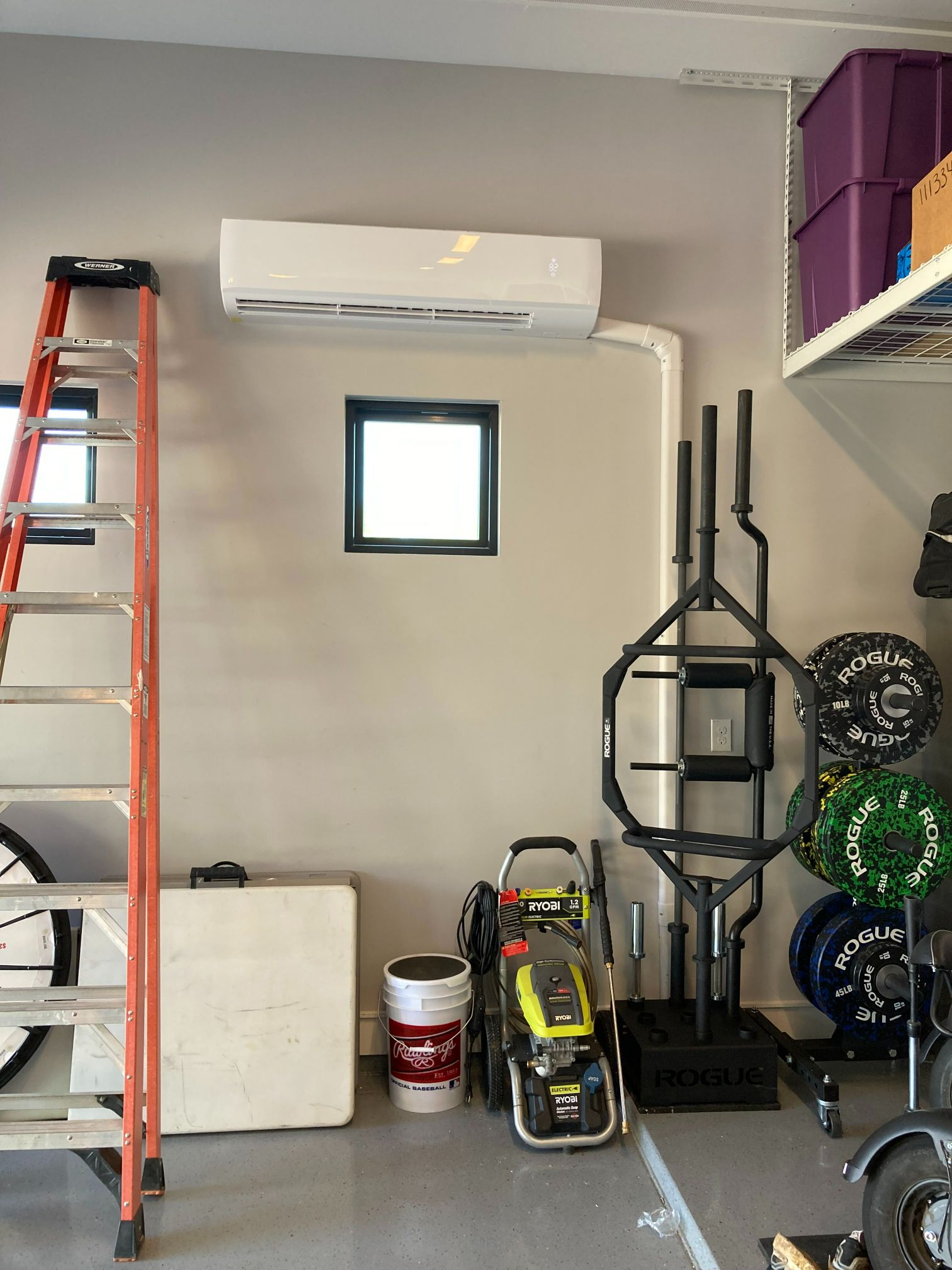 Air conditioning unit above a small window in a garage gym with equipment and a ladder.