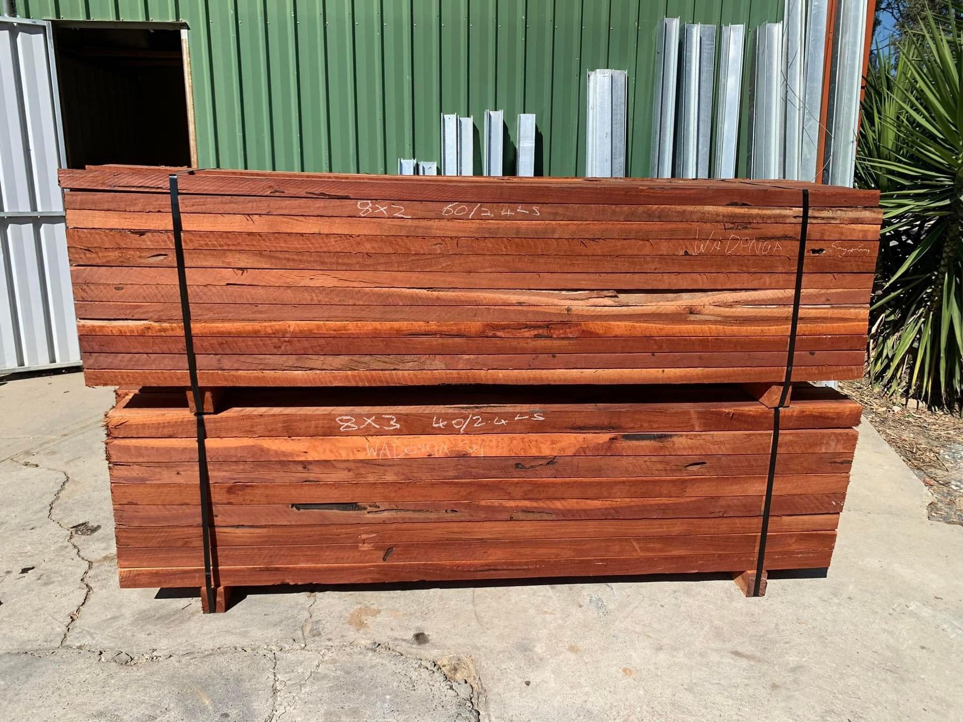 Two stacks of reddish-brown timber planks, bound with black straps, outdoors against a green building. — Wodonga Sand & Soil in Wodonga, VIC
