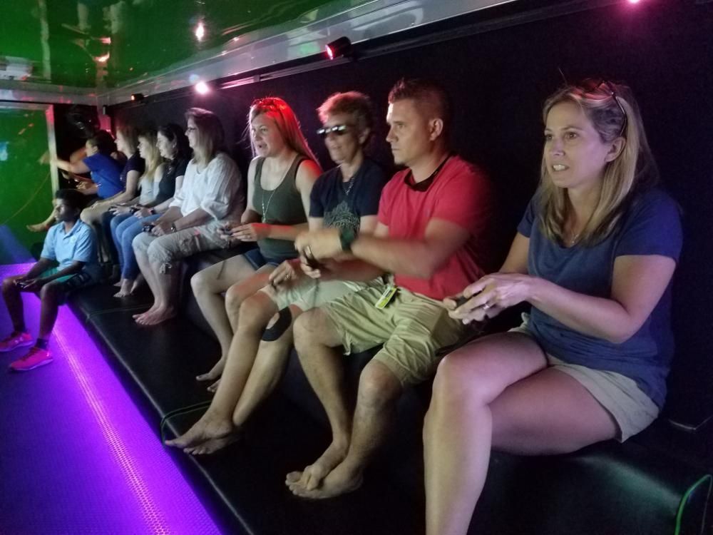 A group of people sit side-by-side in a dim, purple-lit room, looking forward while wearing 3D glasses.