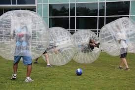 People play bubble soccer on a grassy field in front of a modern building with large glass windows.