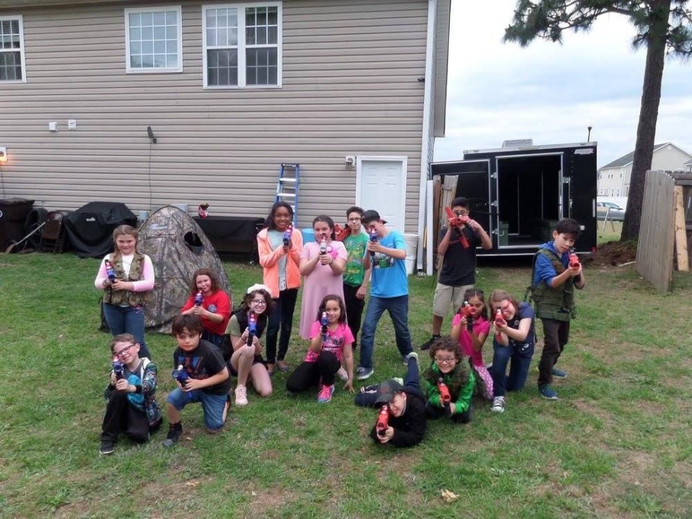 A large group of children and teens posing with toy blasters in a backyard in front of a house and a black trailer.