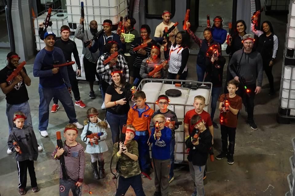 A diverse group of people holding toy laser guns in an indoor arena, posing for a group photo behind a large white container.