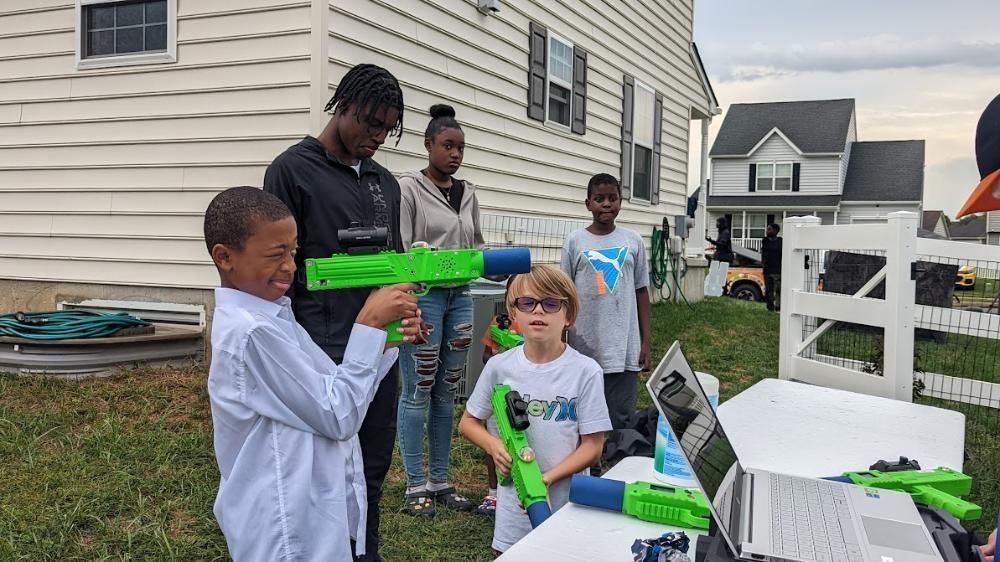 Group standing outdoors, two individuals holding bright green water guns near a table with a laptop.