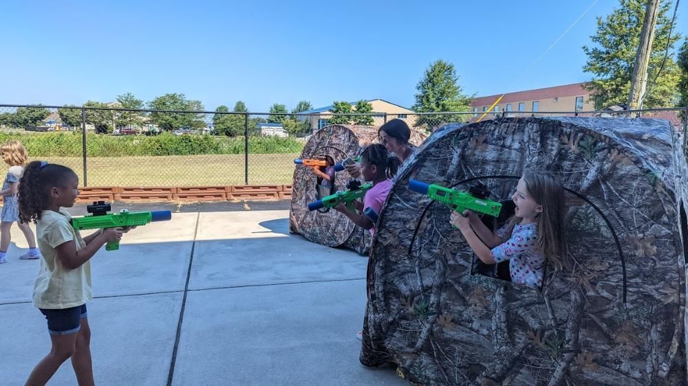 Children playing with toy foam dart blasters outside, ducking behind camouflaged pop-up tents on a sunny day.