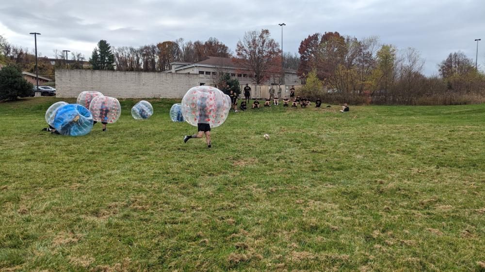 People play bubble soccer on a grassy field, wearing large, transparent inflatable bubbles while running and falling.