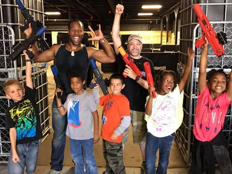 A group of children and adults posing indoors, holding toy laser guns in the air and smiling in a gaming facility.