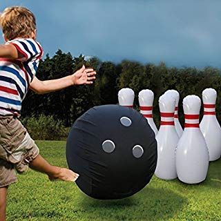 A person kicks a large, black, inflatable bowling ball toward a set of white inflatable pins on a grassy lawn.