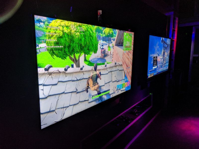 Two wall-mounted televisions display gameplay from Fortnite in a darkened room with ambient purple lighting.