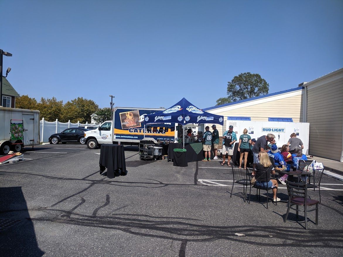People gather around a food truck and tent set up in a parking lot on a sunny day.