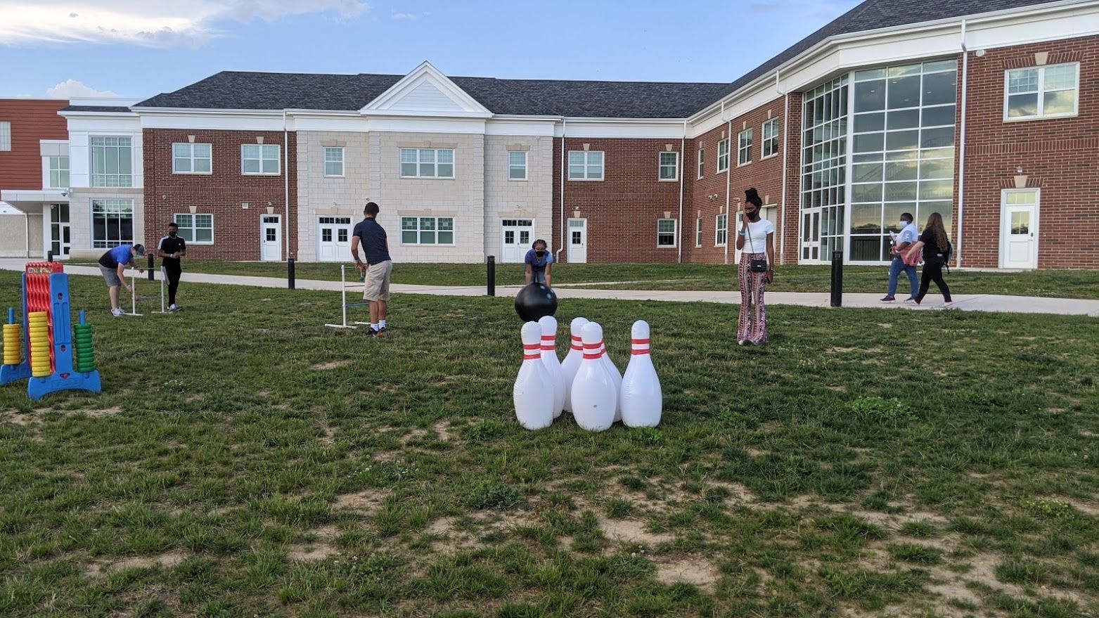 People play with large inflatable bowling pins and yard games on a grassy lawn in front of a brick campus building.