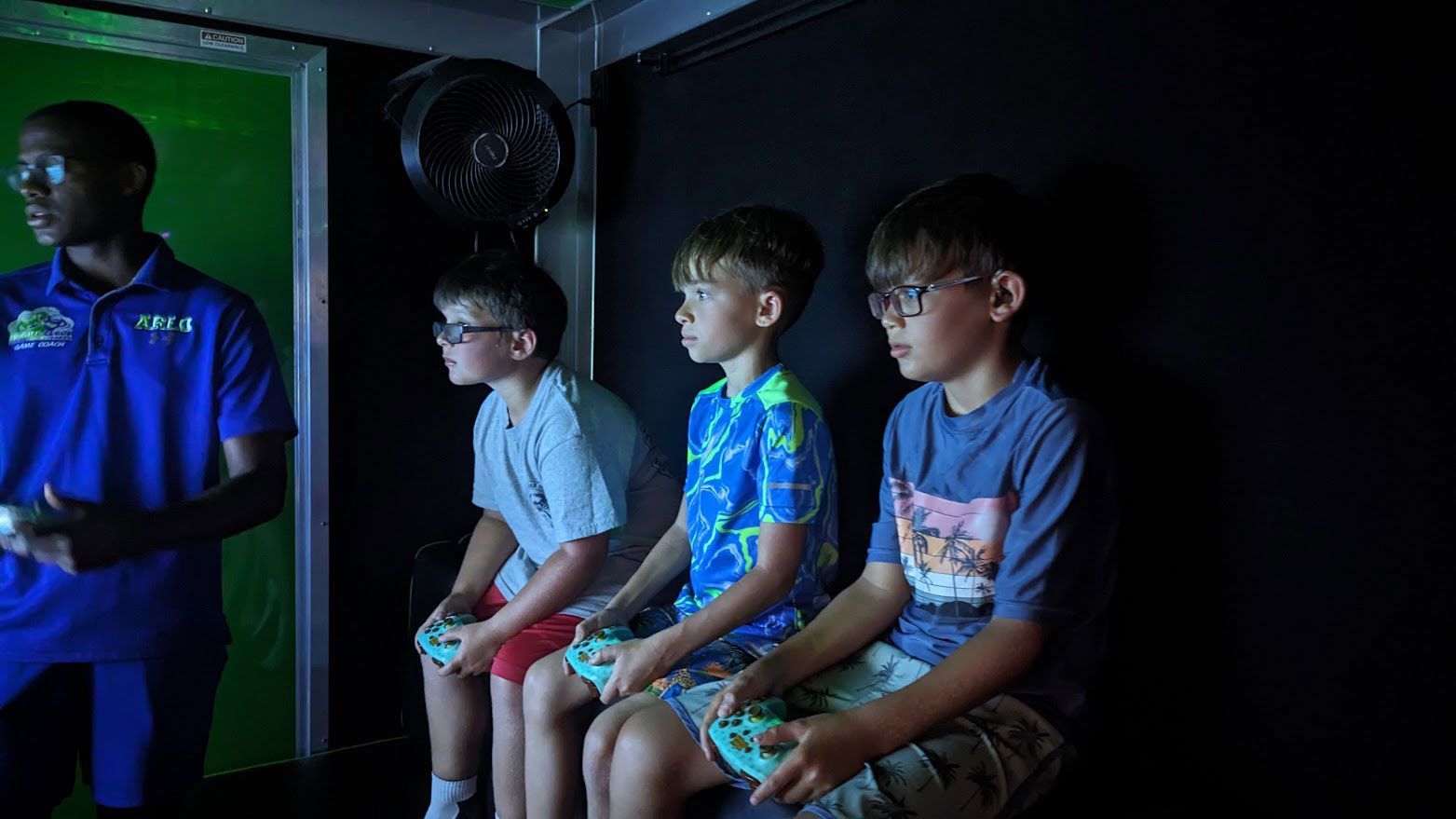 Three people holding game controllers sit on stools in a dim room, facing a screen, while an adult stands to their left.